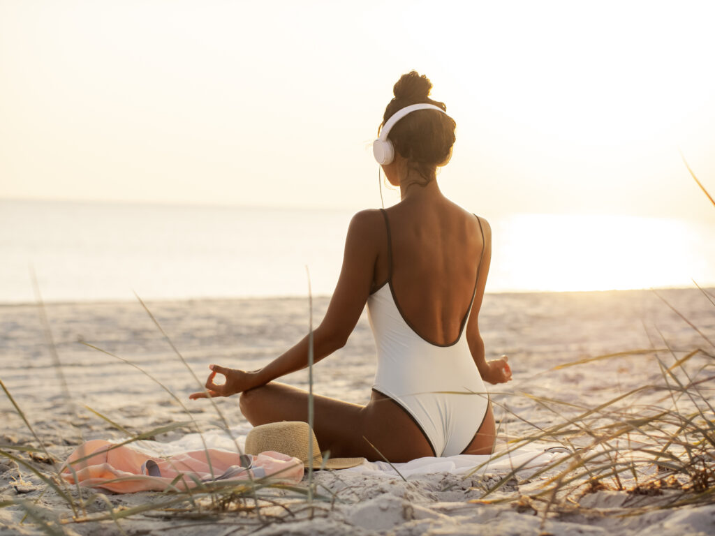 Young beautiful woman in bikini meditating and listening to calm music in yoga lotus pose on deserted tropical beach outdoors. Tanned Mixed race Caucasian Asian slim girl sitting with headphones dressed in swimsuit relaxing listening sounds for relax and meditation at sunset, enjoying her vacations. Rear view